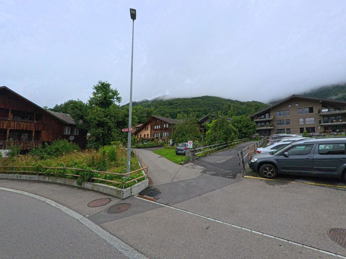 In Hofstetten bei Brienz (3858, Kanton Bern) zweigt ein Fussweg mit Geländer zwischen Wohnhäusern und Parkplätzen ab und führt in Richtung bewaldeter Hänge.