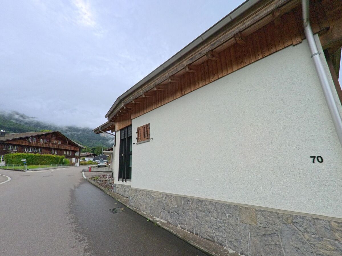 In Hofstetten bei Brienz (3858, Kanton Bern) verläuft eine schmale Strasse an einem hell verputzten Gebäude mit der Hausnummer 70 vorbei, im Hintergrund sind traditionelle Holzhäuser zu sehen.