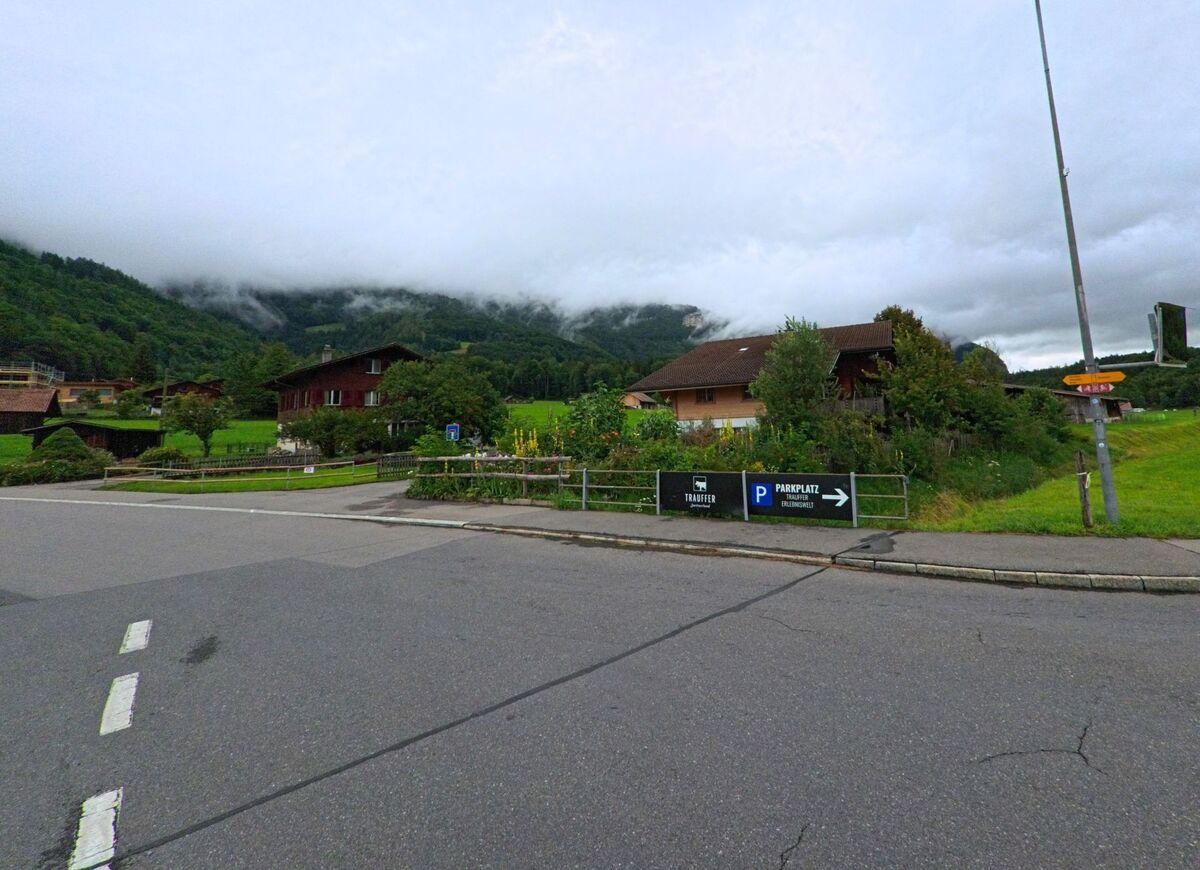 In Hofstetten bei Brienz (3858, Kanton Bern) stehen einzelne Wohnhäuser zwischen Wiesen am Fuss bewaldeter Hügel, mit Hinweisschildern zum Parkplatz und Wanderweg.