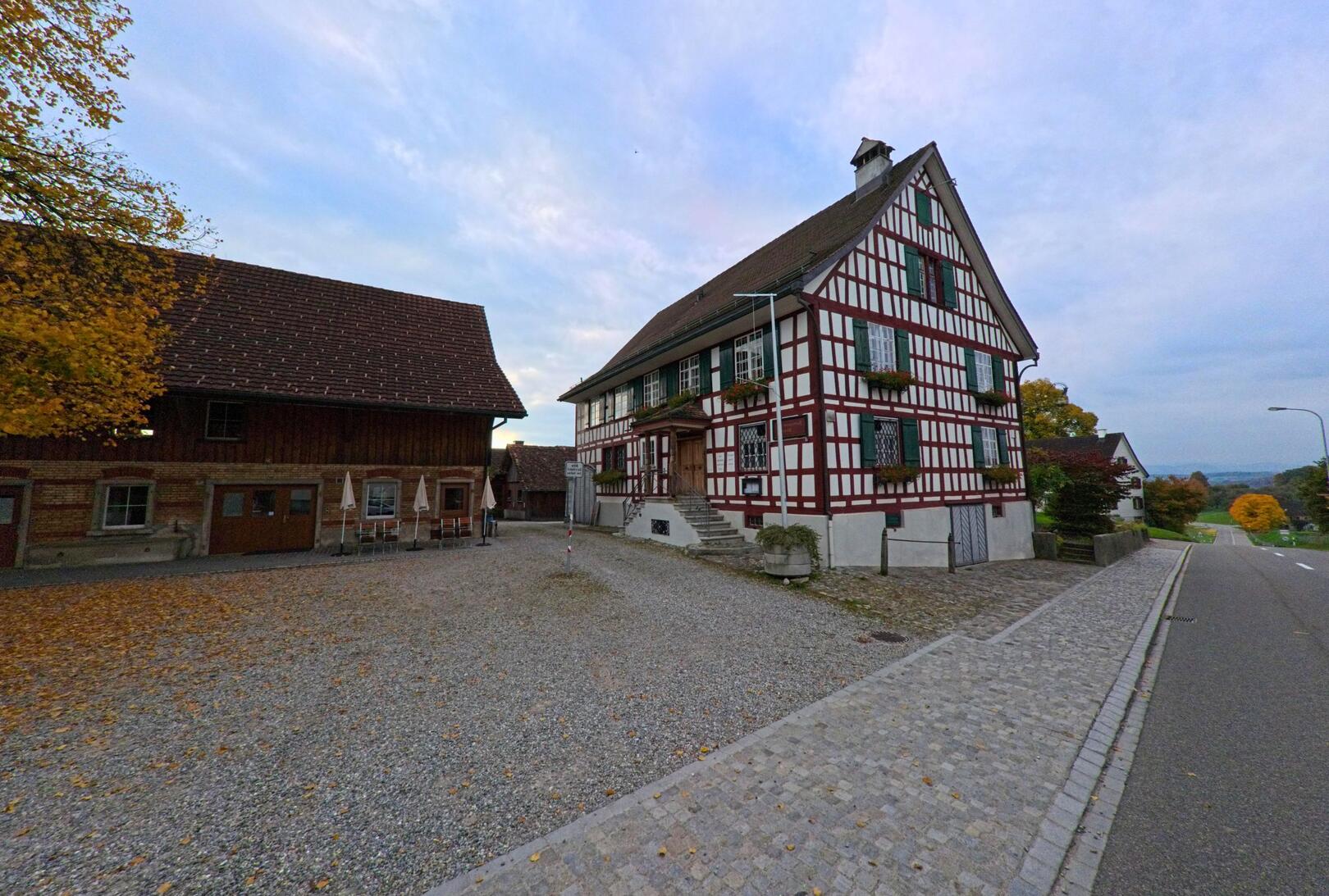 Am Rand einer Strasse steht in Hohentannen (9216, Kanton Thurgau) ein grosses Fachwerkhaus mit Hofplatz und herbstlichen Bäumen.