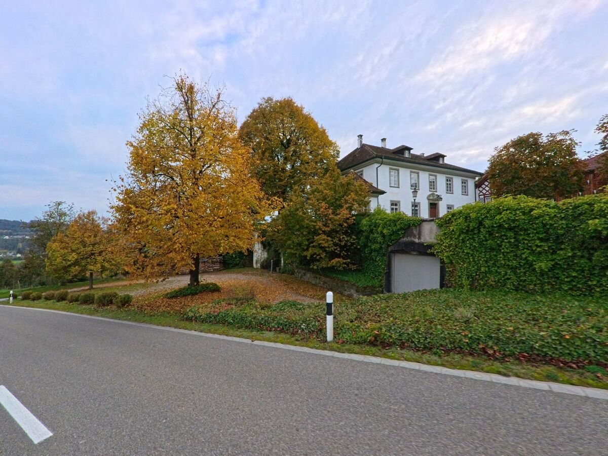 Hinter einer begrünten Mauer liegt in Hohentannen (9216, Kanton Thurgau) ein helles Gebäude zwischen hohen Bäumen mit herbstlichem Laub.