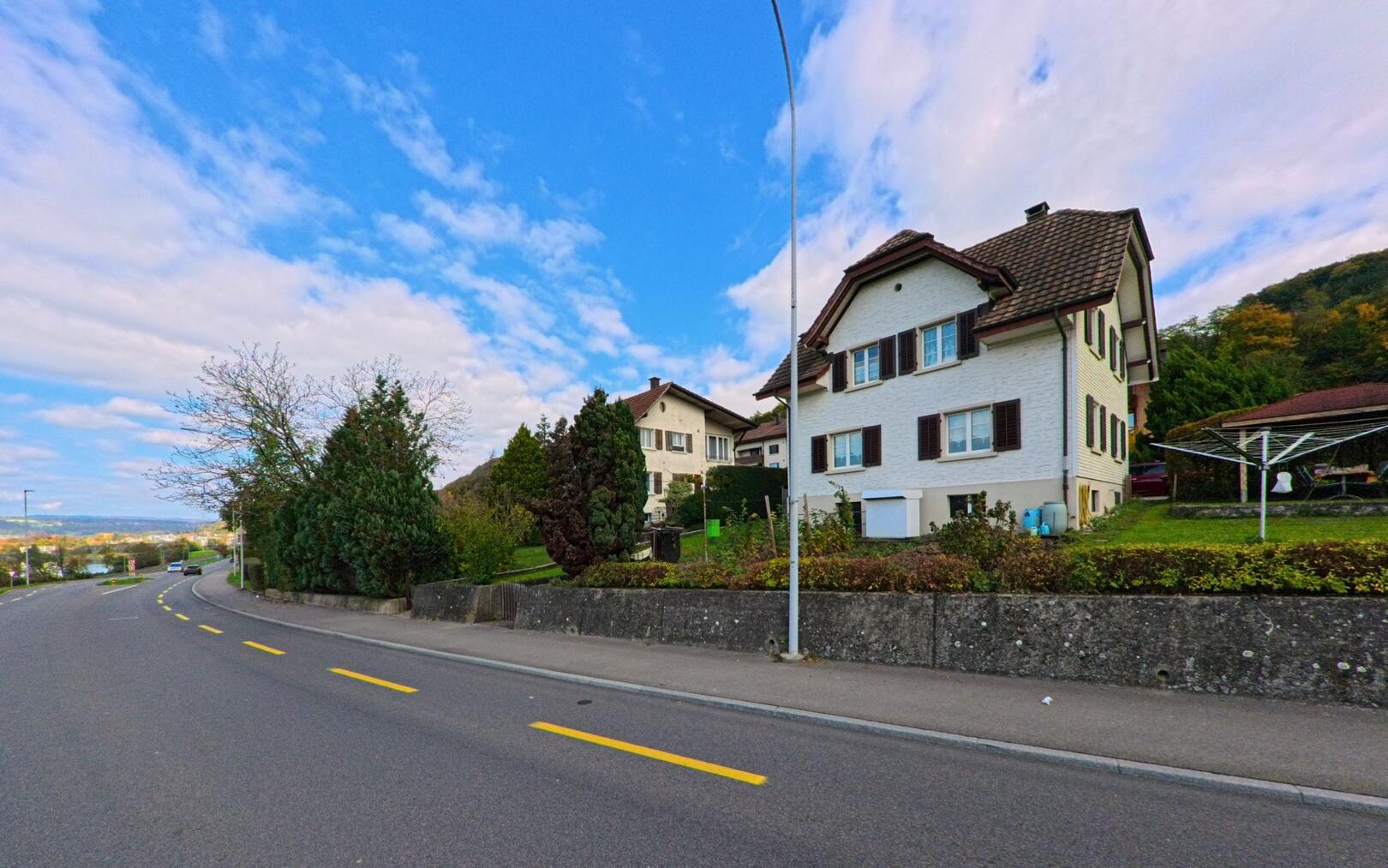 Entlang der Hauptstrasse in Holderbank (5113, Kanton Aargau) stehen gepflegte Einfamilienhaeuser mit Blick auf die umliegenden Huegel.