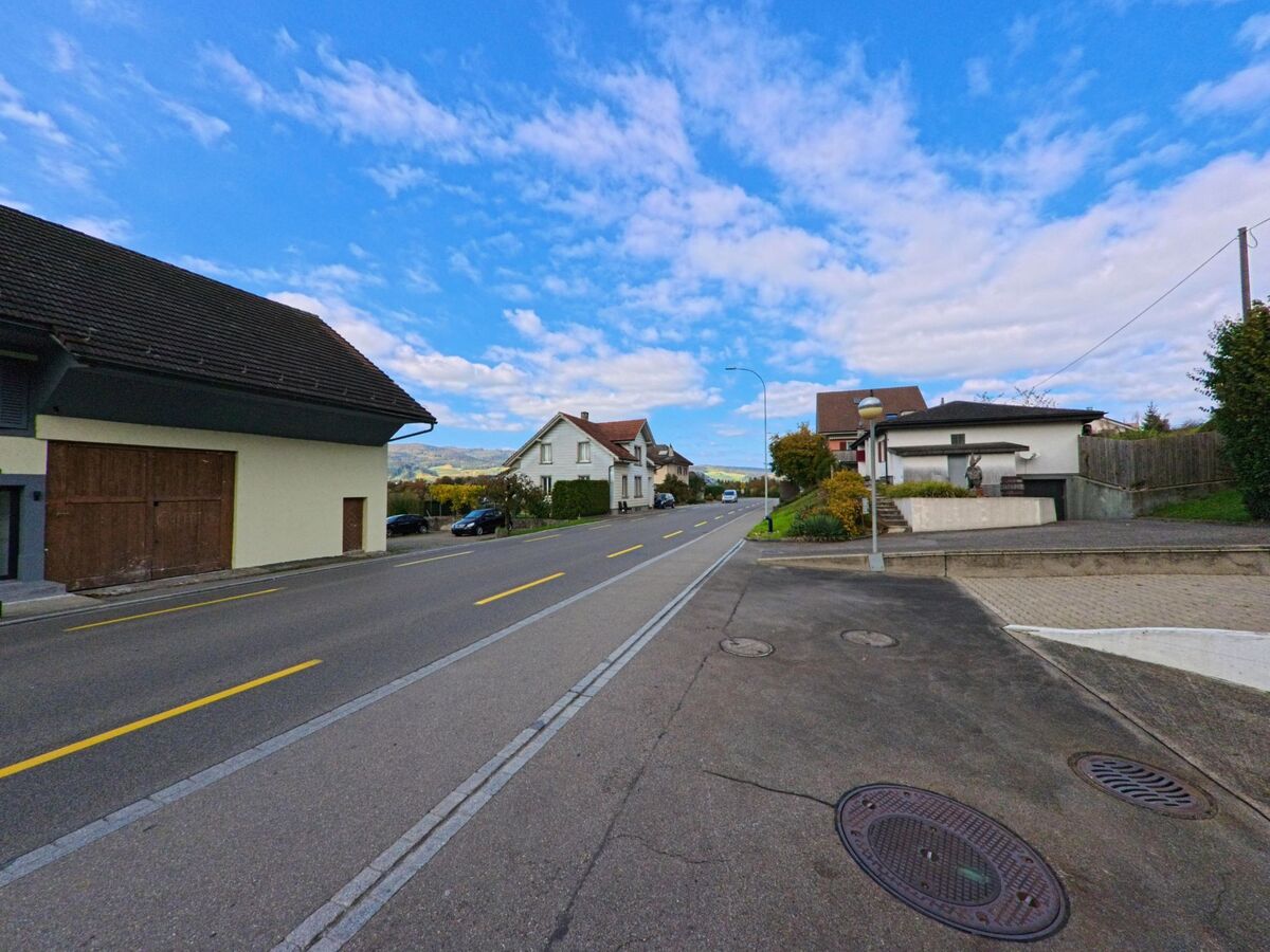 Entlang der Hauptstrasse in Holderbank (5113, Kanton Aargau) stehen mehrere Wohnhaeuser mit gepflegten Vorgartenanlagen unter blauem Himmel.