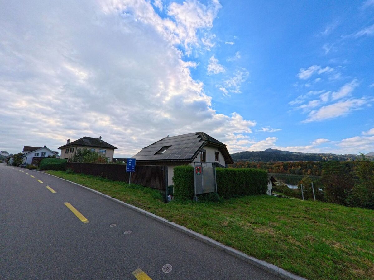 Ein Haus mit Solarpanels auf dem Dach steht am Strassenrand in Holderbank (5113, Kanton Aargau) mit Blick auf die bewaldeten Huegel im Hintergrund.
