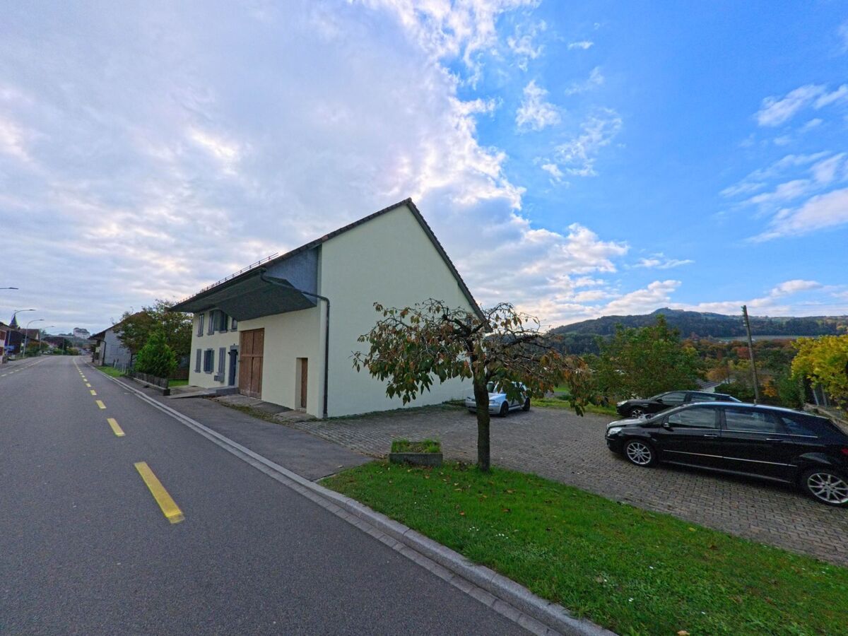 Ein helles Wohnhaus mit Holztoren steht an einer ruhigen Strasse in Holderbank (5113, Kanton Aargau) mit Blick auf die bewaldeten Huegel im Hintergrund.
