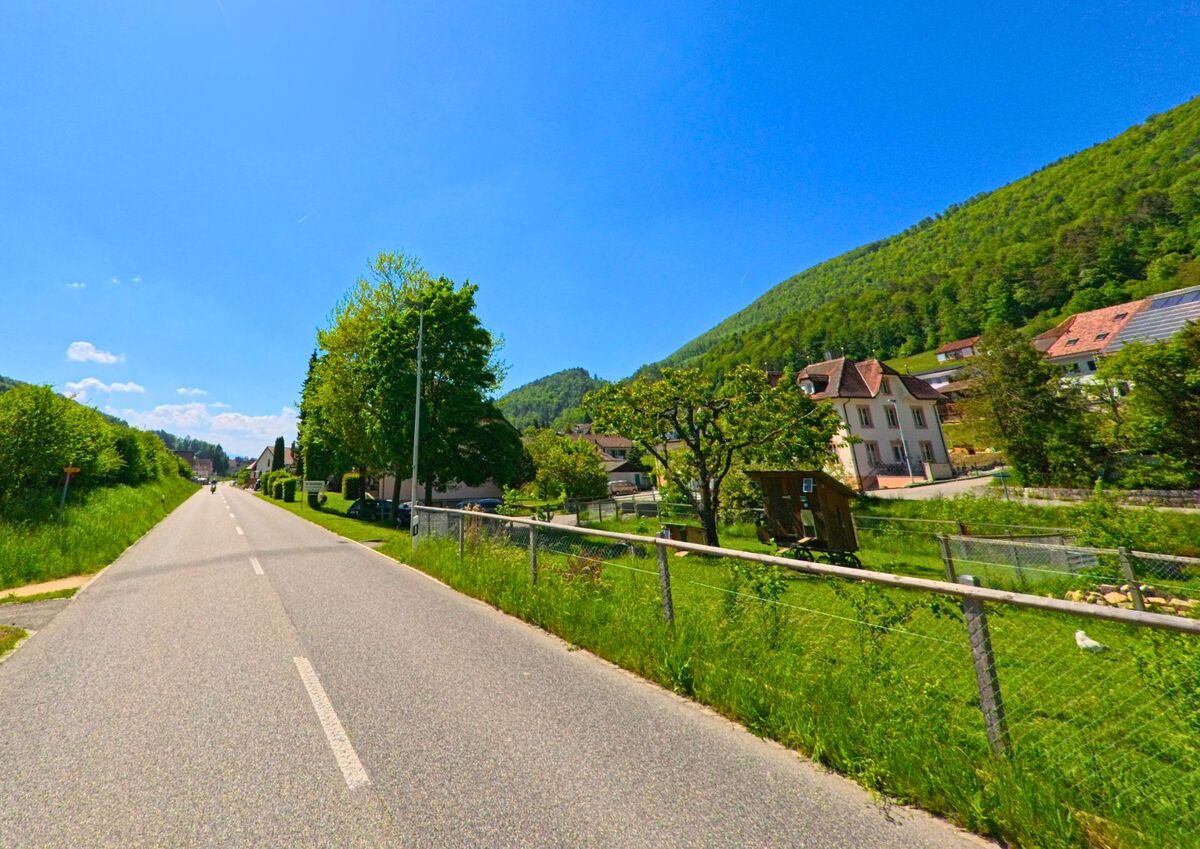 In Holderbank (PLZ 4718, Kanton Solothurn) verlaeuft eine laendliche Strasse zwischen Huegellandschaft und kleinen Hausgaerten mit Ausblick auf traditionelle Wohnhaeuser.