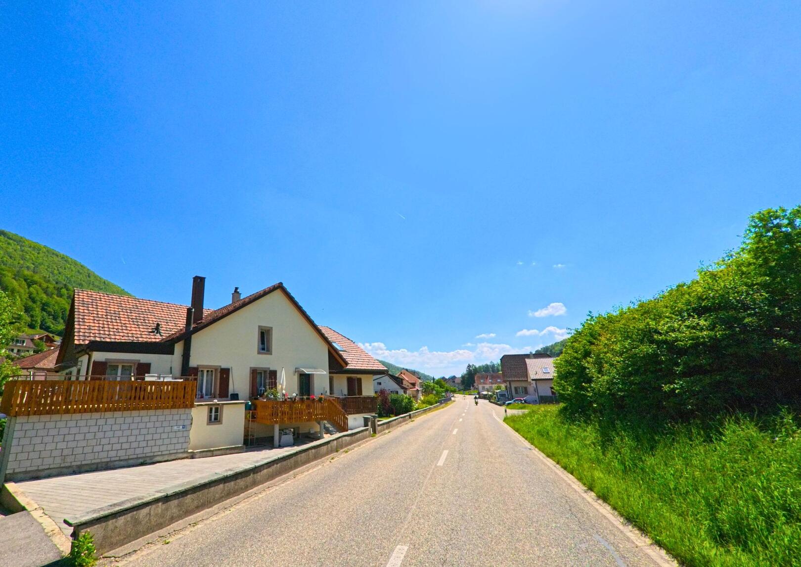 In Holderbank (4718, Kanton Solothurn) verlaeuft eine Hauptstrasse unter strahlender Sonne vorbei an Wohnhaeusern am Hang eines bewaldeten Huegels.