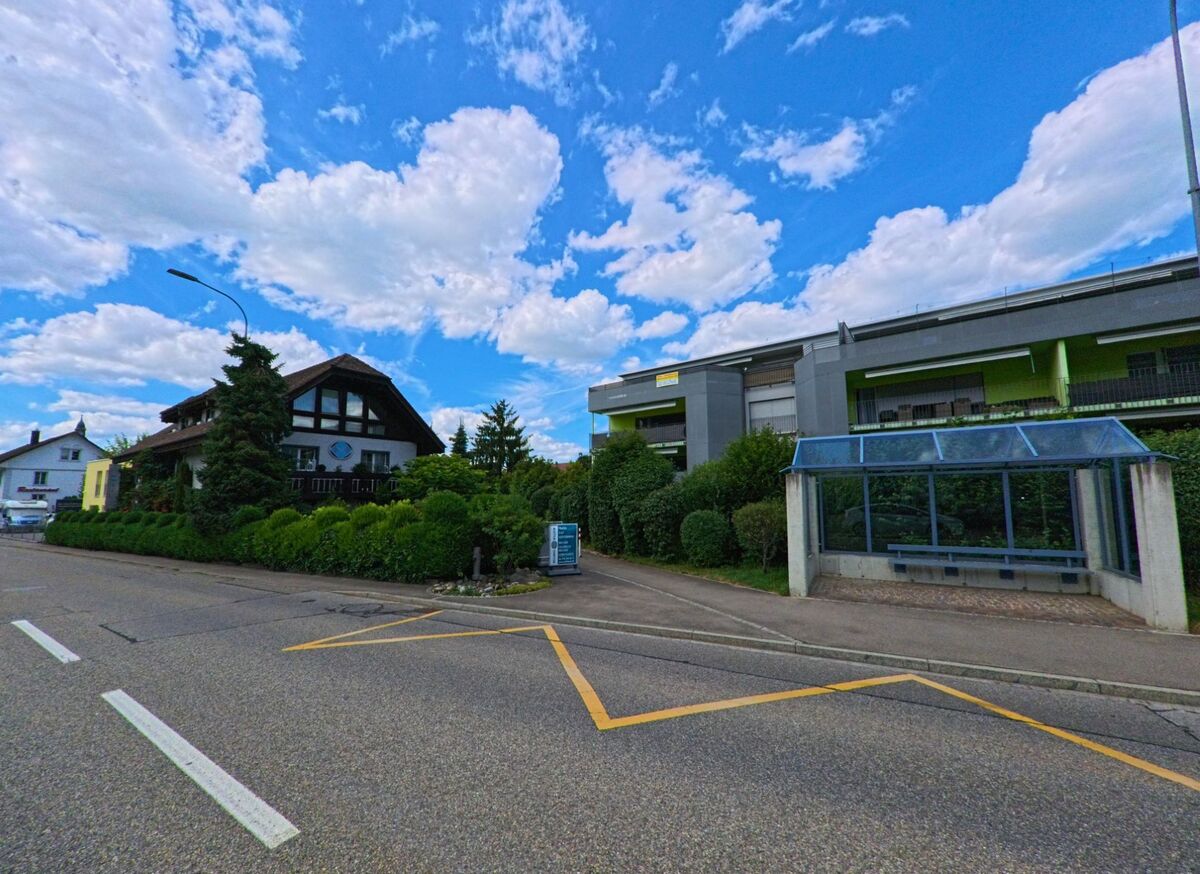 In Holziken (5043, Kanton Aargau) liegt eine Bushaltestelle mit Glasdach gegenueber einem traditionellen Haus und einem modernen Wohnbau mit gruener Fassade.