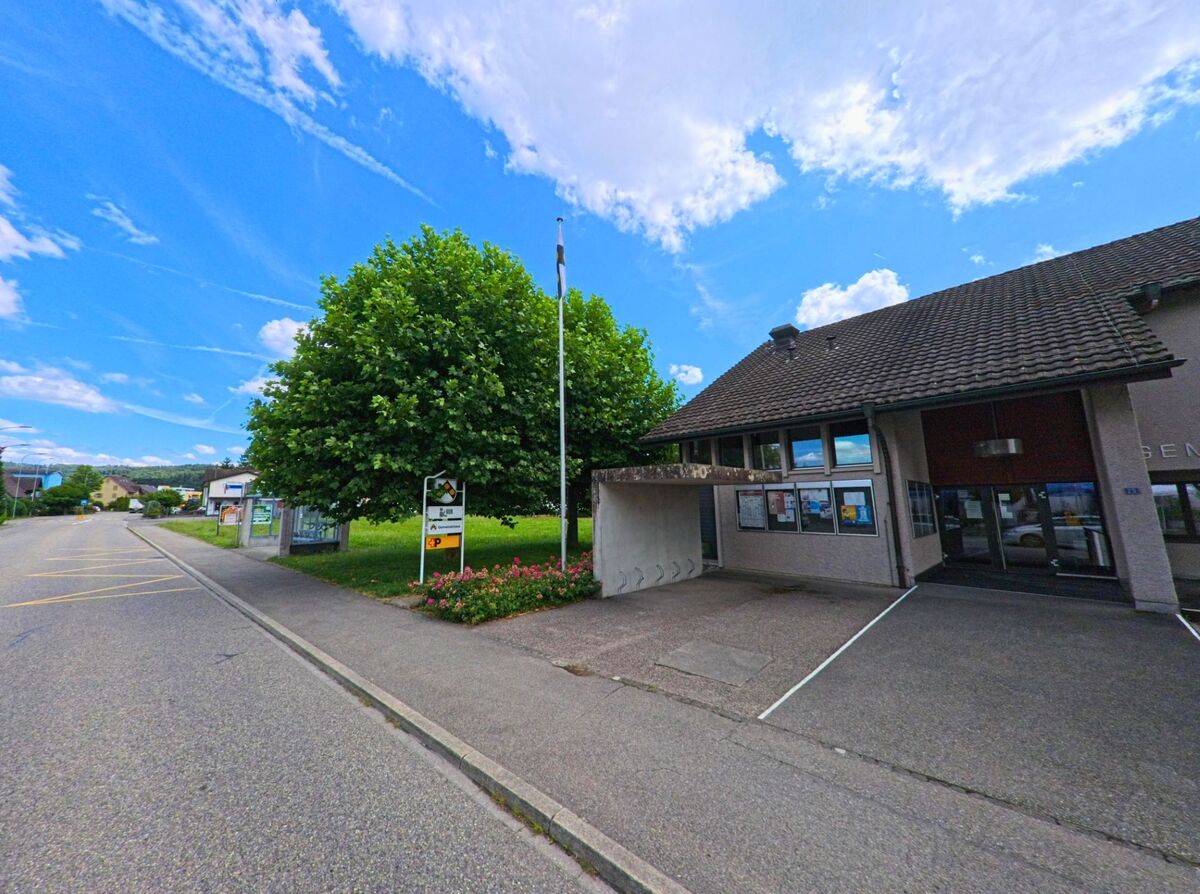 Das Gemeindehaus von Holziken (5043, Kanton Aargau) liegt an einer ruhigen Strasse mit Fahrradstaendern und Informationstafeln vor einem grossen Baum.