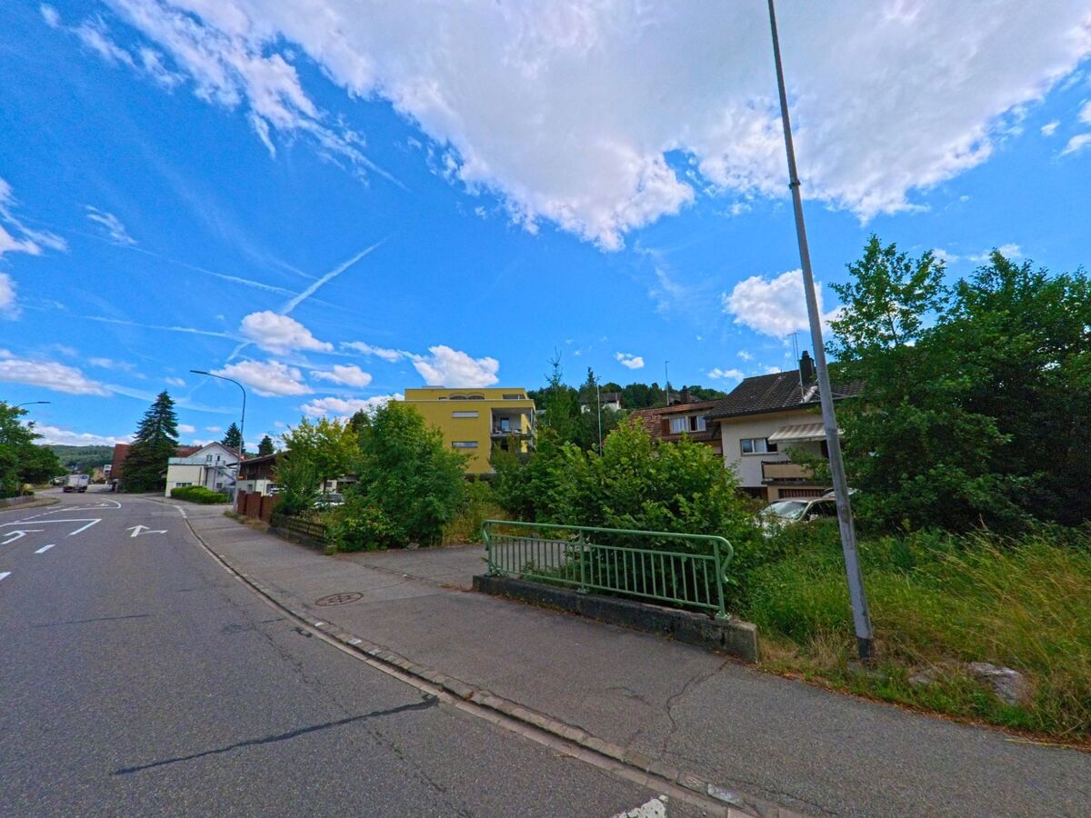 In Holziken (5043, Kanton Aargau) fuehrt eine Strasse mit Abbiegespuren an einem kleinen Brueckengeländer vorbei, dahinter liegen Wohnhaeuser und dichter Bewuchs.