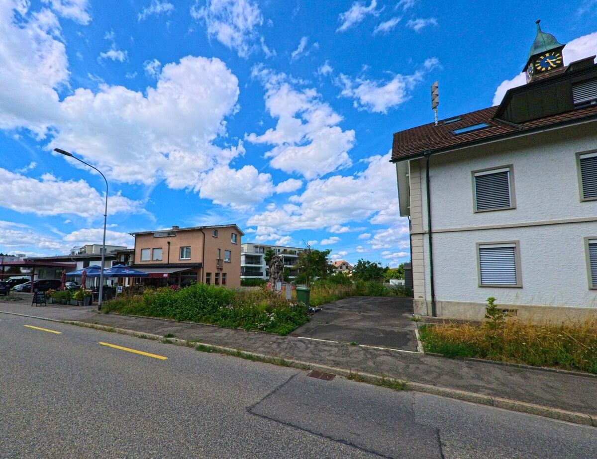 In Holziken (5043, Kanton Aargau) steht ein Gebaeude mit markantem Uhrturm neben einer asphaltierten Einfahrt, gegenueber befindet sich ein kleines Restaurant mit Terrasse.
