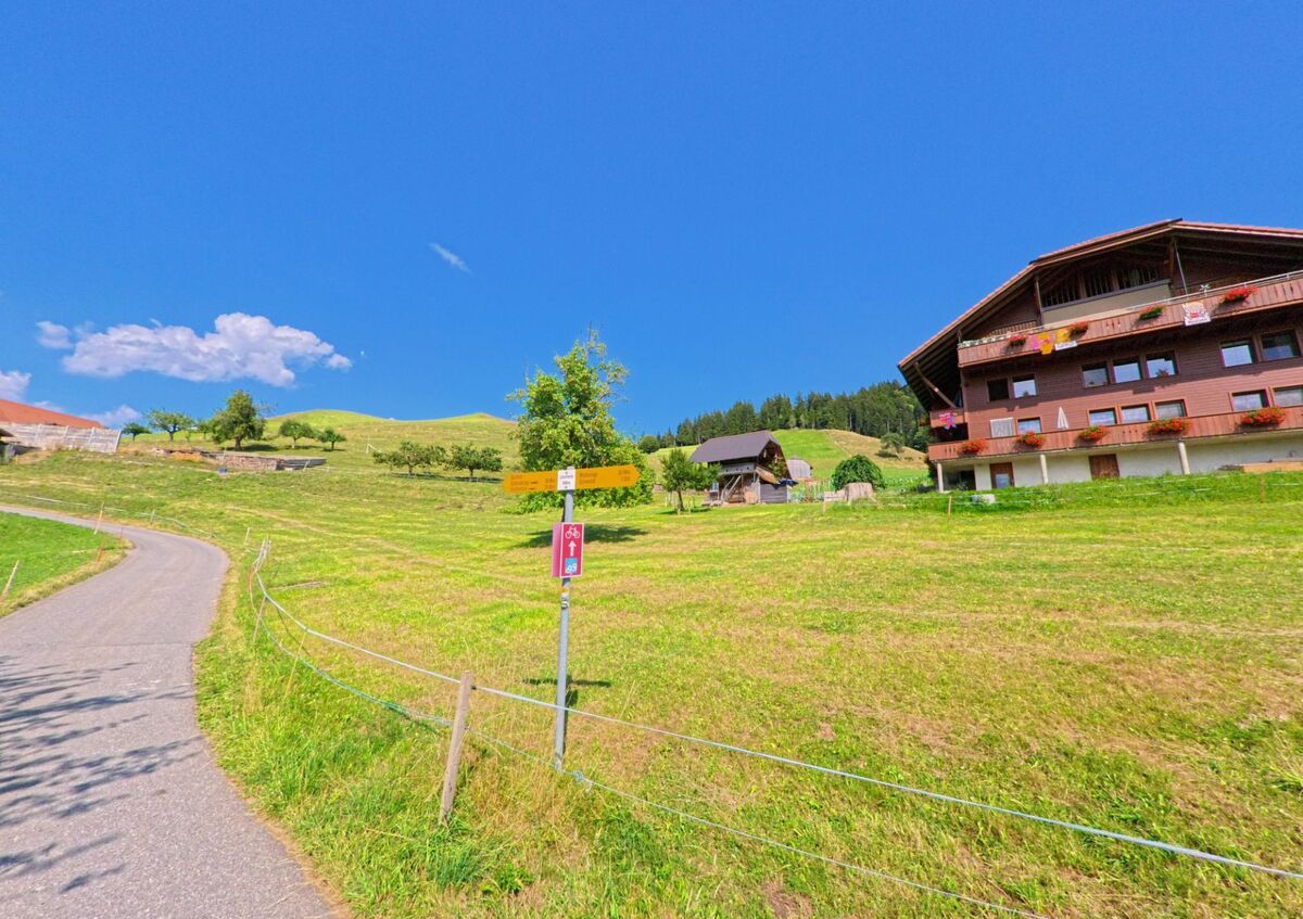 In Homberg (3622, Kanton Bern) steht ein braunes Wohnhaus mit Balkonen an einem sonnigen Hang, daneben ein gelber Wanderwegweiser und ein Veloweg-Schild vor gruener Kulisse.