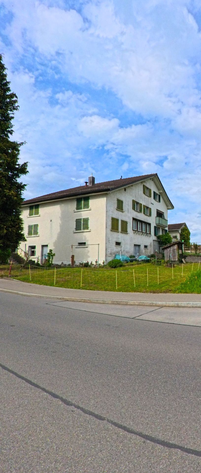Ein aelteres Wohnhaus mit grünen Fensterlaeden steht in Hombrechtikon (8634, Kanton Zürich) leicht erhoeht an einer Strasse, umgeben von einem einfachen Vorgarten.