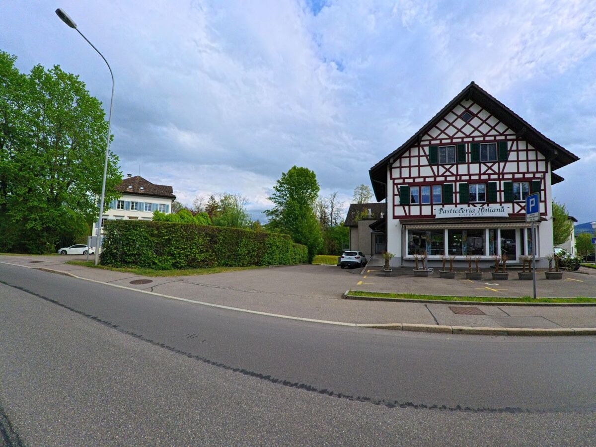 Ein traditionelles Fachwerkgebaeude in Hombrechtikon (8634, Kanton Zürich) steht an einer ruhigen Strasse, flankiert von dichter Begruenung und weiteren Wohnhaeusern.