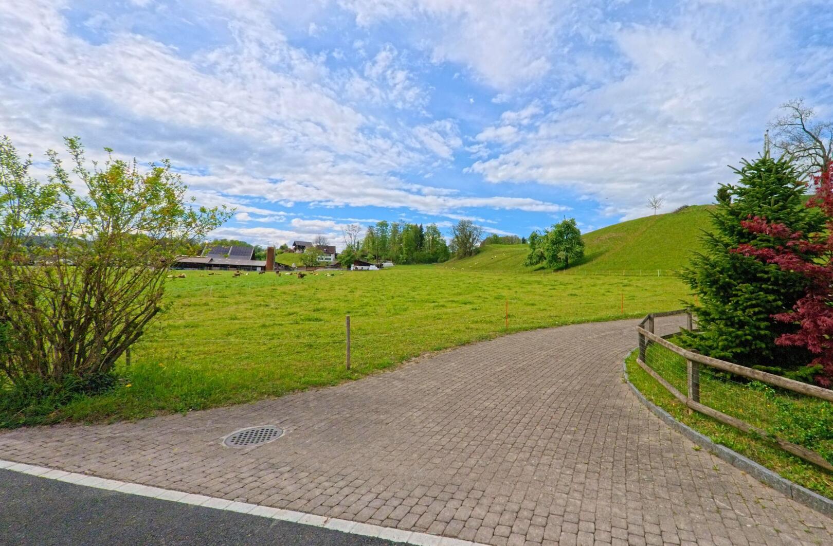 In Hombrechtikon (8634, Kanton Zürich) führt ein gepflasterter Weg entlang einer grünen Wiese, mit einigen Häusern und Hügeln im Hintergrund unter einem klaren Himmel.