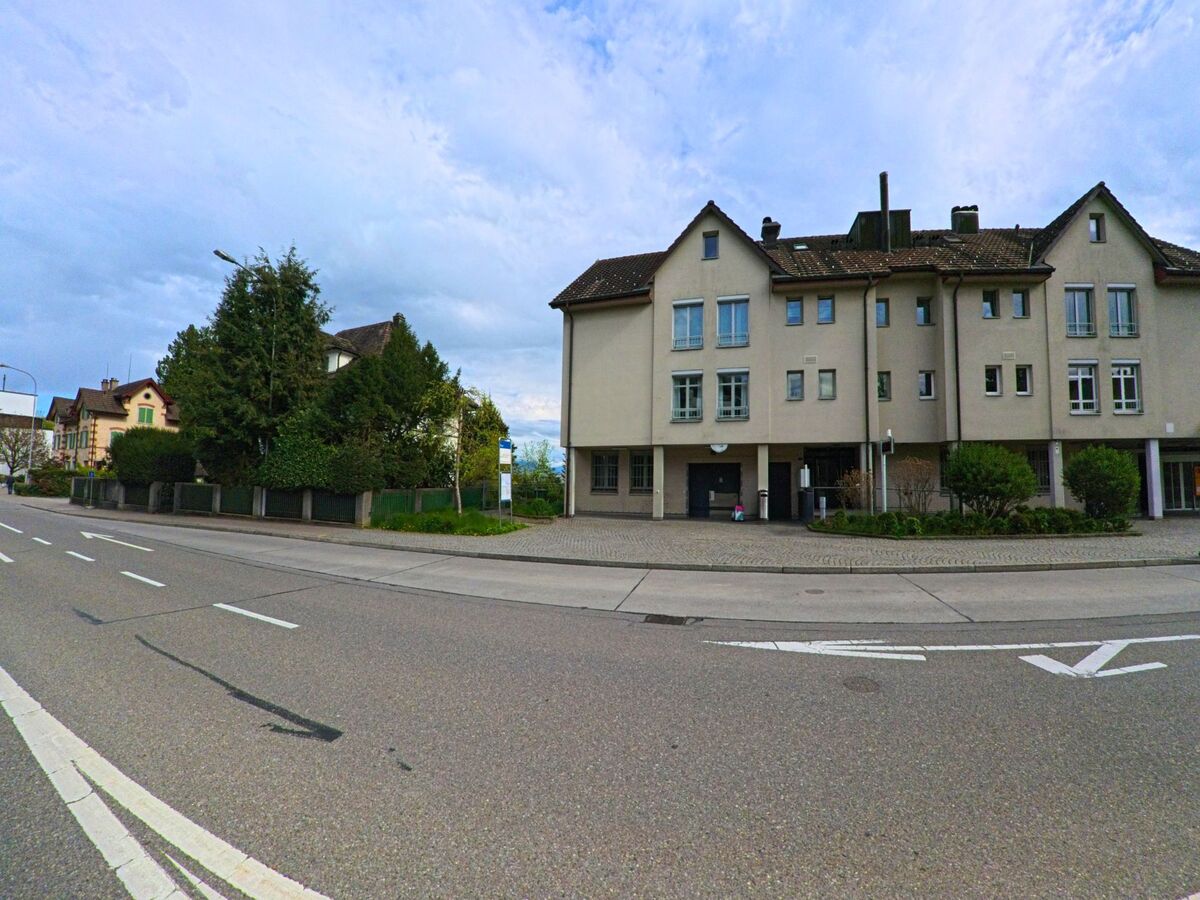 Entlang der Hauptstrasse in Hombrechtikon (8634, Kanton Zürich) stehen mehrgeschossige Wohnhaeuser neben dicht bepflanzten Gartengrundstuecken und einer Bushaltestelle.
