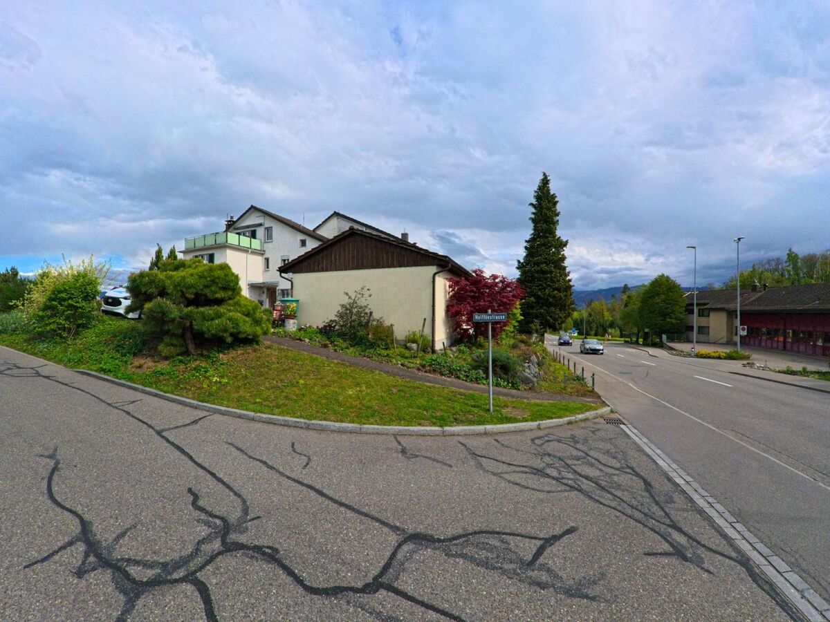 An einer Strassenkreuzung in Hombrechtikon (8634, Kanton Zürich) liegen begruente Vorgartenbereiche und Wohnhaeuser, waehrend Autos die leicht abfallende Hauptstrasse passieren.