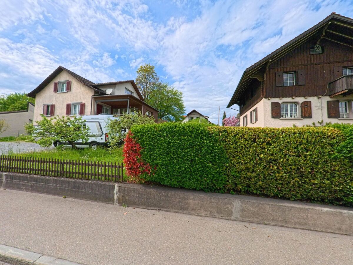Zwei traditionelle Wohnhaeuser in Hombrechtikon (8634, Kanton Zürich) stehen hinter einem gepflegten Garten mit dichter Hecke, Baumblueten und einem geparkten Fahrzeug.
