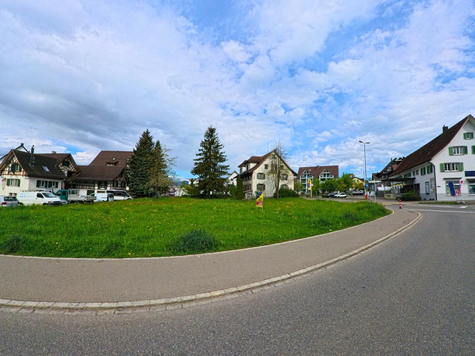 Alt-Text: Eine begruente Verkehrsinsel in Hombrechtikon (8634, Kanton Zürich) liegt zwischen traditionellen Wohnhaeusern und einem leicht belebten Strassenabschnitt.