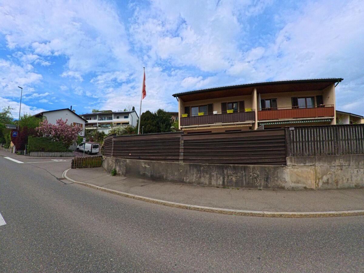 Entlang einer ruhig verlaufenden Strasse in Hombrechtikon (8634, Kanton Zürich) stehen mehrere Wohnhaeuser, ein blühender Baum und eine abgegrenzte Terrasse mit Holzsichtschutz.