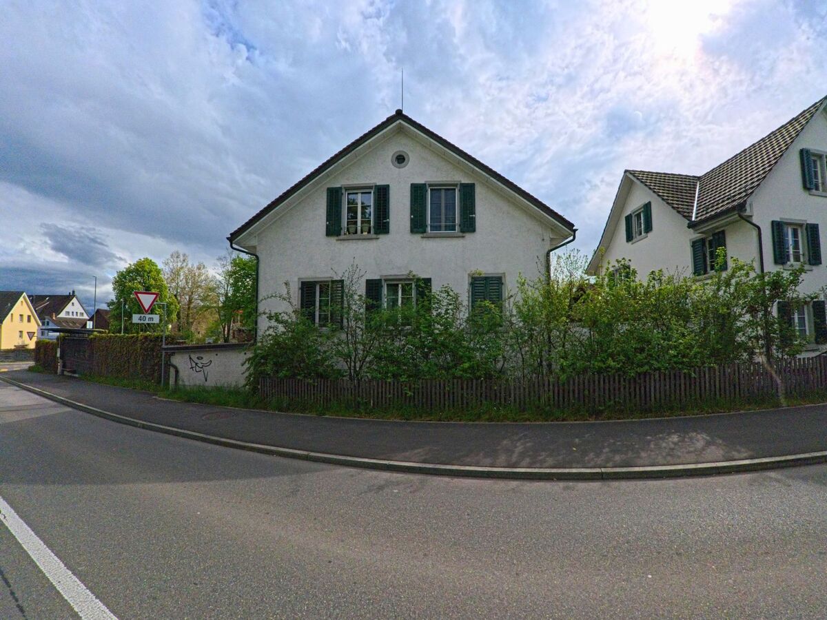 Ein weisses Wohnhaus mit gruenen Fensterlaeden steht in Hombrechtikon (8634, Kanton Zürich) hinter einer dichten Hecke an einer abgerundeten Strassenkurve.