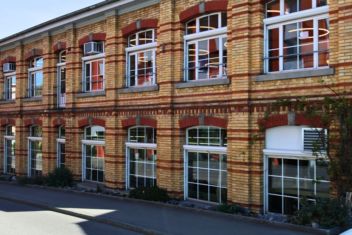  In Horgen (8810, Kanton Zürich) erstreckt sich eine ehemalige Industriehalle mit gelb-roter Ziegelfassade und grossen Fenstern entlang eines schmalen Gehwegs.