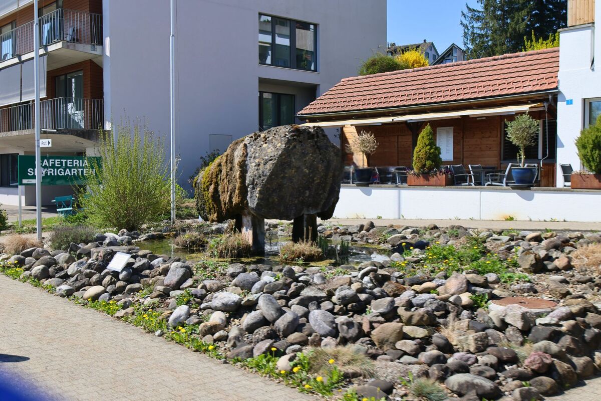  In Horgen (8810, Kanton Zürich) steht ein moosbedeckter Felsblock auf Stelzen als Kunstobjekt in einem steinernen Garten vor einer Cafeteria.