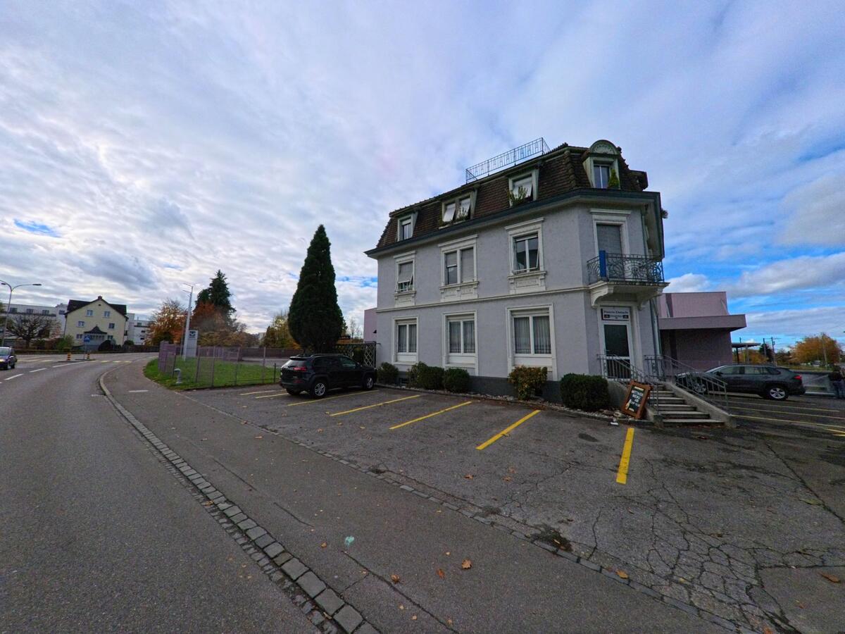 Vor einem grauen Gebaeude mit Balkon und Parkplaetzen liegt eine Seitenflaeche an der Strasse in Horn (9326, Kanton Thurgau) unter wolkigem Himmel.