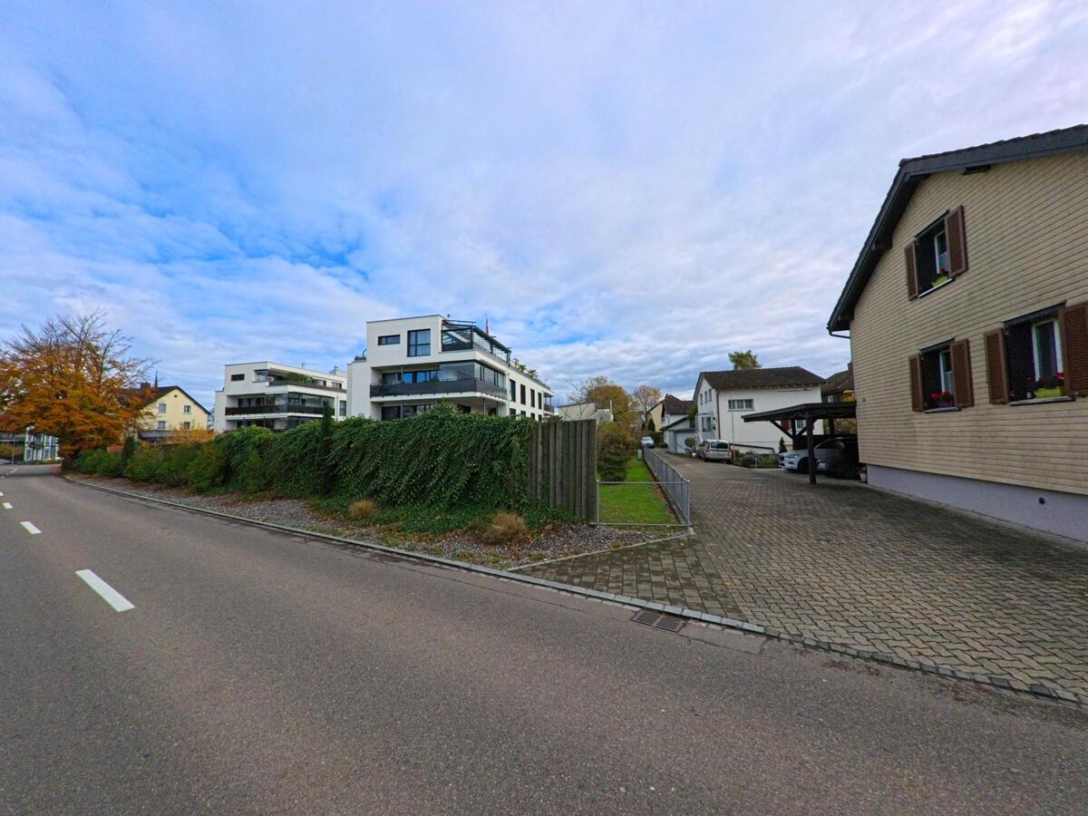 Am Rand einer Wohnstrasse in Horn (9326, Kanton Thurgau) stehen moderne Mehrfamilienhaeuser hinter einer begrünten Hecke neben Einfamilienhaeusern.