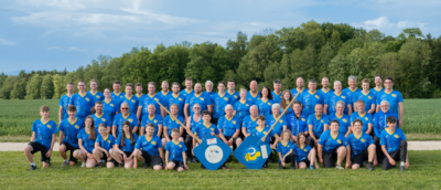 Auf einer Wiese in Fraubrunnen (3317, Kanton Bern) steht eine grosse Gruppe von Personen in einheitlichen Shirts fuer ein gemeinsames Gruppenfoto.