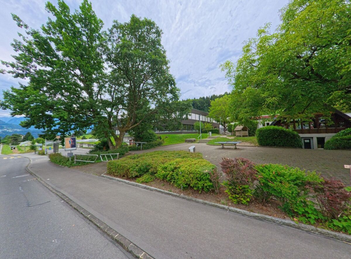 In Horw (6047, Kanton Luzern) erstreckt sich eine grosszuegige Schulanlage mit Spielplatz und Sitzgelegenheiten unter schattenspendenden Baeumen am Strassenrand.