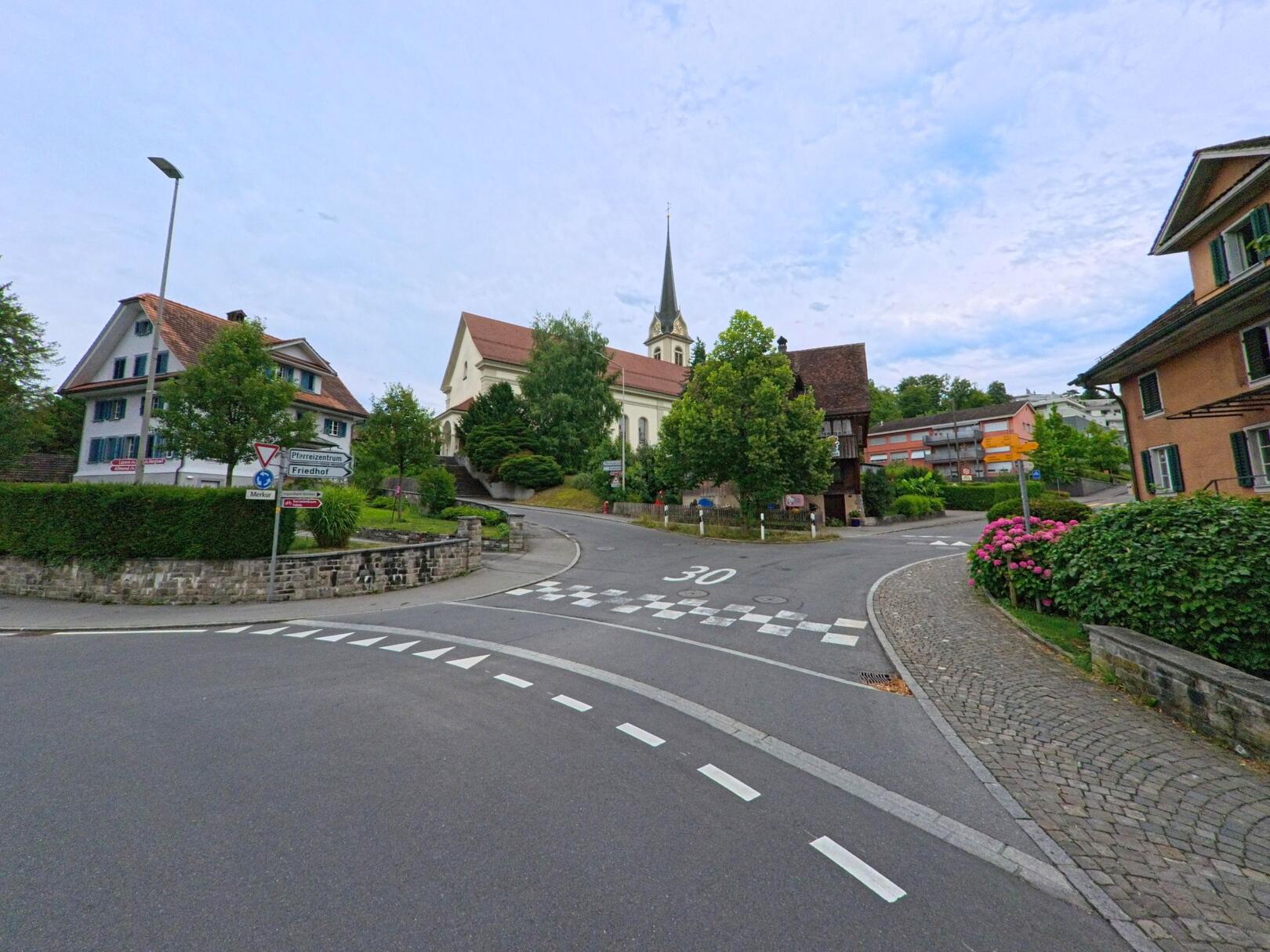 Im Zentrum von Horw (6048, Kanton Luzern) fuehrt eine verkehrsberuhigte Strasse zur Kirche mit spitzem Turm, umrahmt von traditionellen Wohnhaeusern und Bluetengewaechsen.