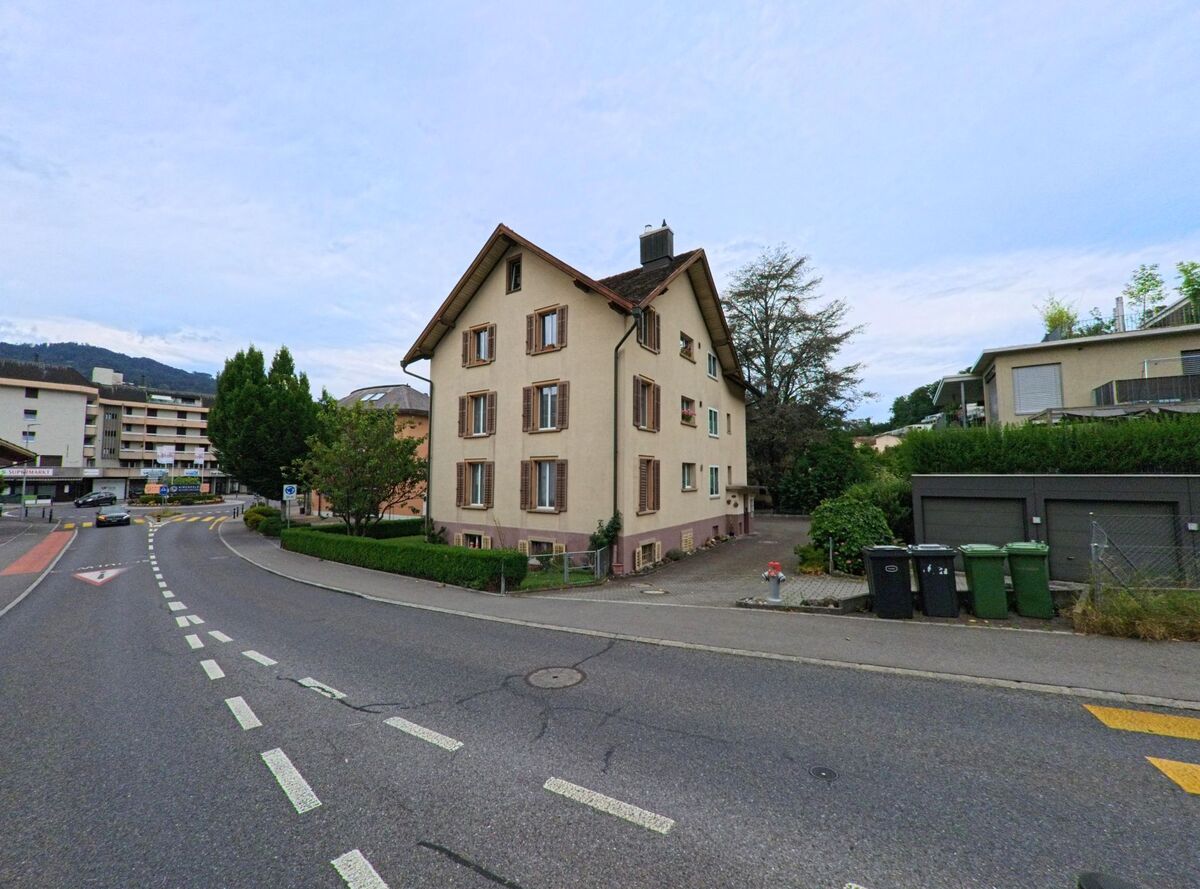 Ein Mehrfamilienhaus mit Fensterlaeden steht in Horw (6048, Kanton Luzern) an einer breiten Strassenkreuzung, umgeben von moderner Bebauung und Bergkulisse im Hintergrund.
