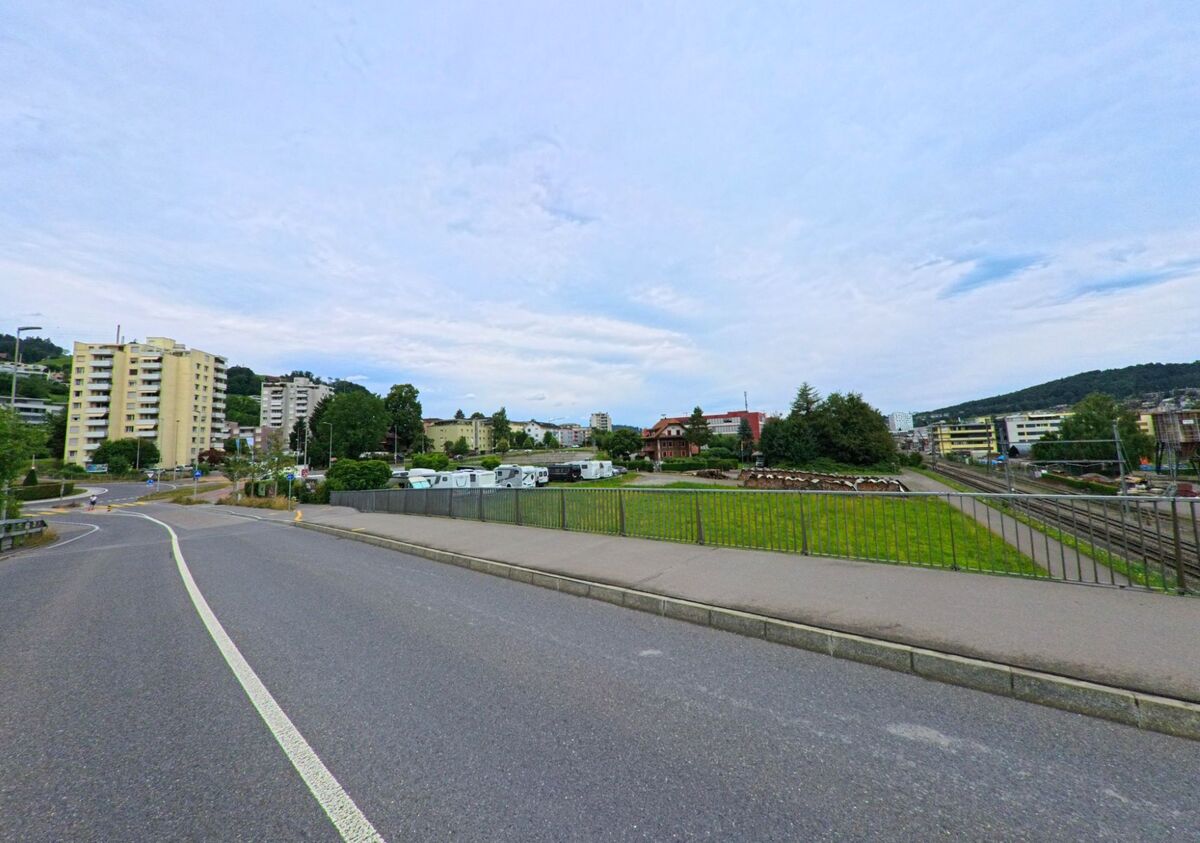 In Horw (6048, Kanton Luzern) fuehrt eine Strasse mit angrenzendem Fussweg durch ein urbanes Gebiet mit Hochhaeusern, Gruenflaechen und Bahntrassee.