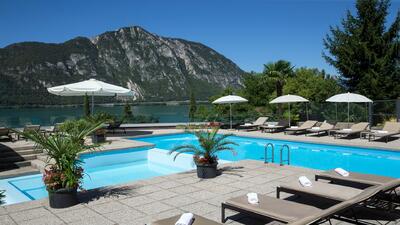 Vom Poolbereich des Hotel Campione in Bissone (6816, Kanton Tessin) öffnet sich ein weiter Blick über den Luganersee und die umliegenden Berge unter blauem Himmel.
