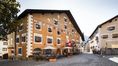 Das Hotel Crusch Alva in Zuoz (7524, Kanton Graubuenden) beeindruckt mit seiner historischen Fassade, Wappenmalereien und einer Terrasse mit Sonnenschirmen.