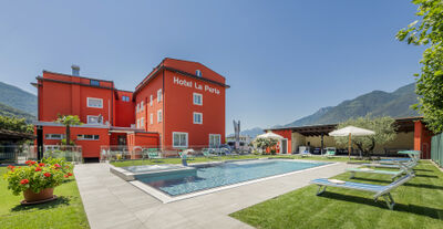 Das Hotel La Perla in S. Antonino (6592, Kanton Tessin) präsentiert sich mit leuchtend roter Fassade, gepflegtem Garten und Aussenpool vor der Kulisse der Tessiner Berge.