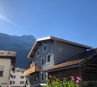 In Andeer (7440, Kanton Graubuenden) steht das Hotel Pitschonia mit grauer Fassade und Holzbalkon vor dem Hintergrund bewaldeter Berghaenge.