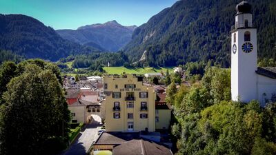 In Andeer (7440, Kanton Graubuenden) steht das Hotel Restaurant Fravi mit gelber Fassade im Dorfzentrum, umgeben von Bergen und einer Kirche mit markantem Turm.