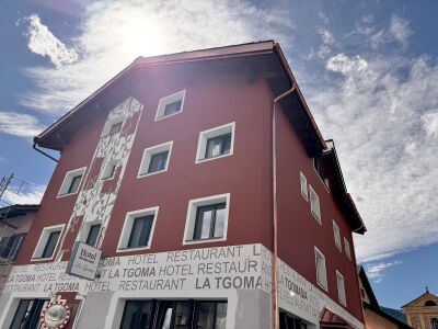 In Lantsch/Lenz (7083, Kanton Graubuenden) steht das rot gestrichene Hotel Restaurant La Tgoma mit weissem Fassadenmuster unter einem sonnigen Himmel.