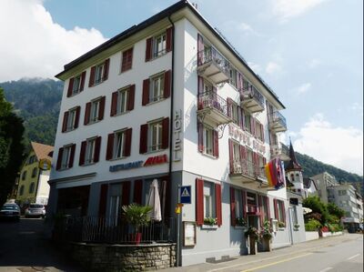 Das Hotel Rigi in Vitznau (6354, Kanton Luzern) steht an einer Strasse mit roten Fensterläden und Balkonen vor einer Bergkulisse.