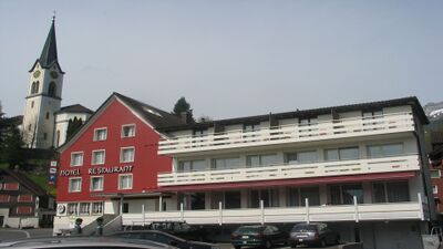 Das Hotel Schäfli in Gams (9473, Kanton St. Gallen) praesentationiert sich mit rotem Haupthaus und weissem Anbau, im Hintergrund ist die Dorfkirche sichtbar.