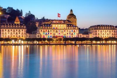 Das Hotel Schweizerhof in Luzern (6002, Kanton Luzern) spiegelt sich abends beleuchtet im ruhigen Wasser des Vierwaldstaettersees.