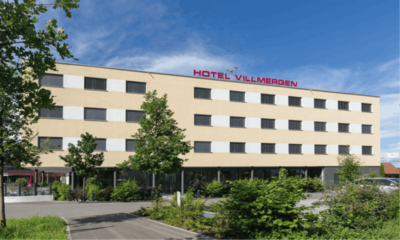 Vor dem modernen Hotel Villmergen in Villmergen (5612, Kanton Aargau) stehen junge Baeume und gepflegte Straucher unter blauem Himmel.