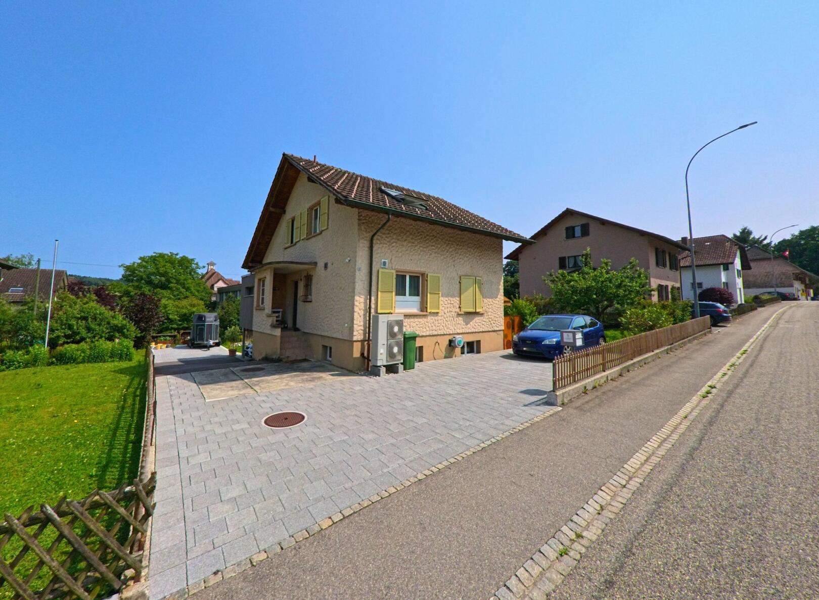 Ein Einfamilienhaus mit hellen Fensterlaeden steht in Hubersdorf (4535, Kanton Solothurn) an einer gepflasterten Einfahrt mit geparktem Auto.