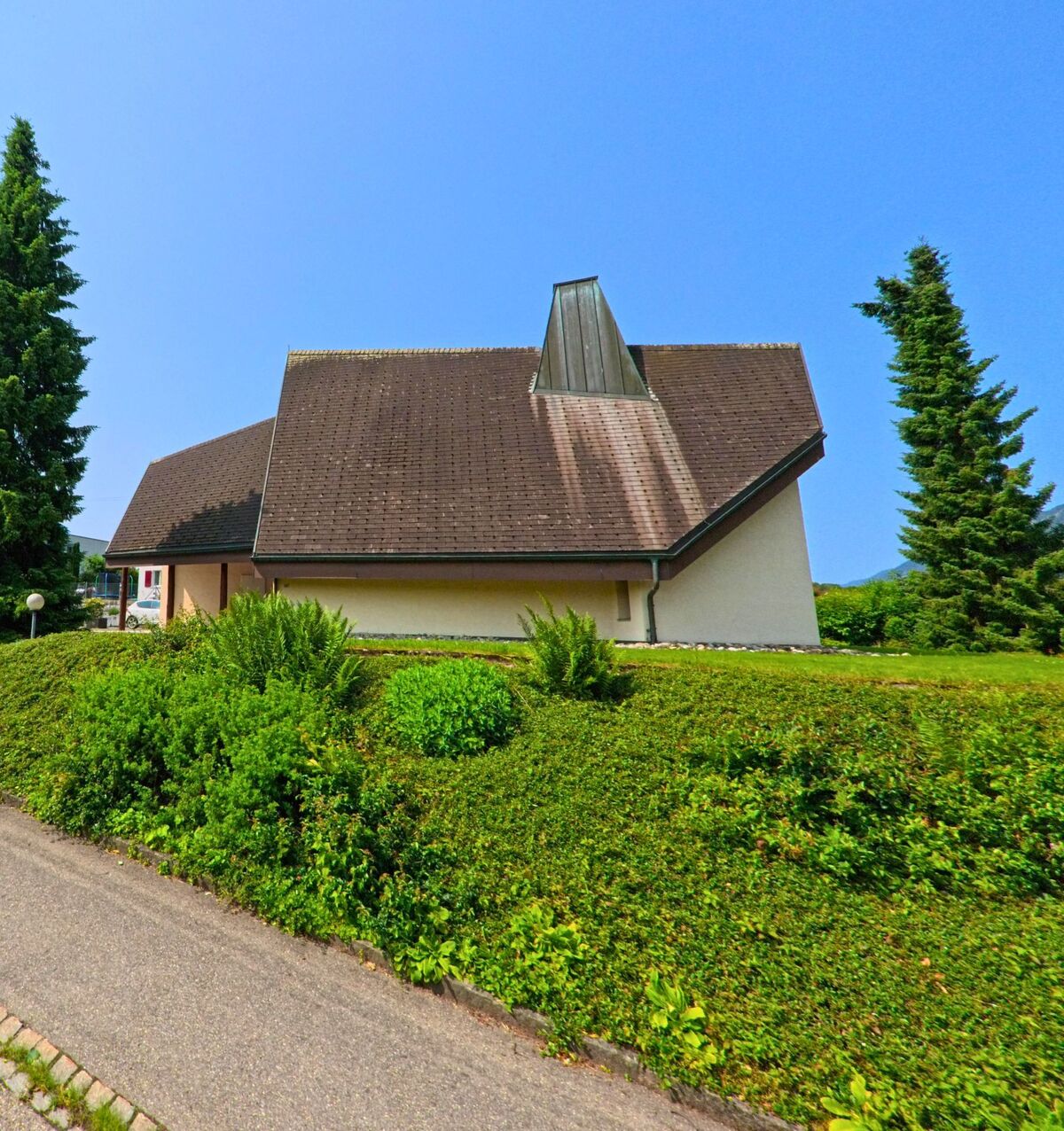 Eine Kirche mit markantem Dach und hohem Aufsatz steht in Hubersdorf (4535, Kanton Solothurn) hinter einer gepflegten Hecke.