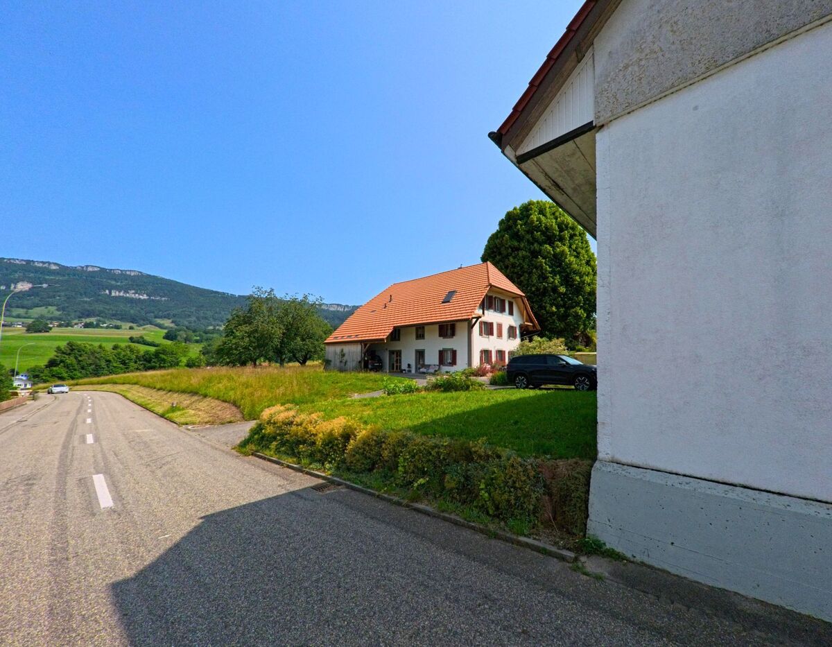 Ein Wohnhaus mit roten Fensterlaeden liegt in Hubersdorf (4535, Kanton Solothurn) an einer Landstrasse mit Blick auf bewaldete Huegel.