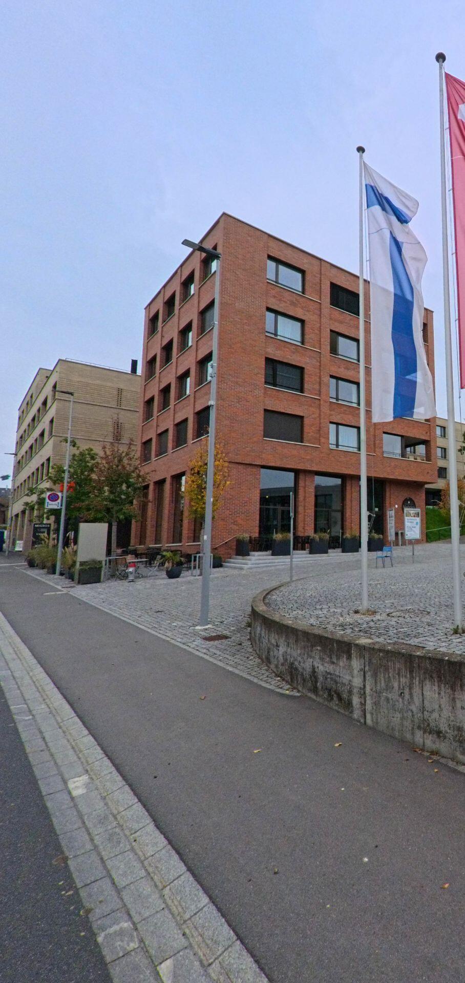 Ein Backsteingebaeude in Hueenenberg (6331, Kanton Zug) steht an einem gepflasterten Vorplatz mit Fahnenmasten und kleinen Sitzbereichen entlang des Gehwegs.