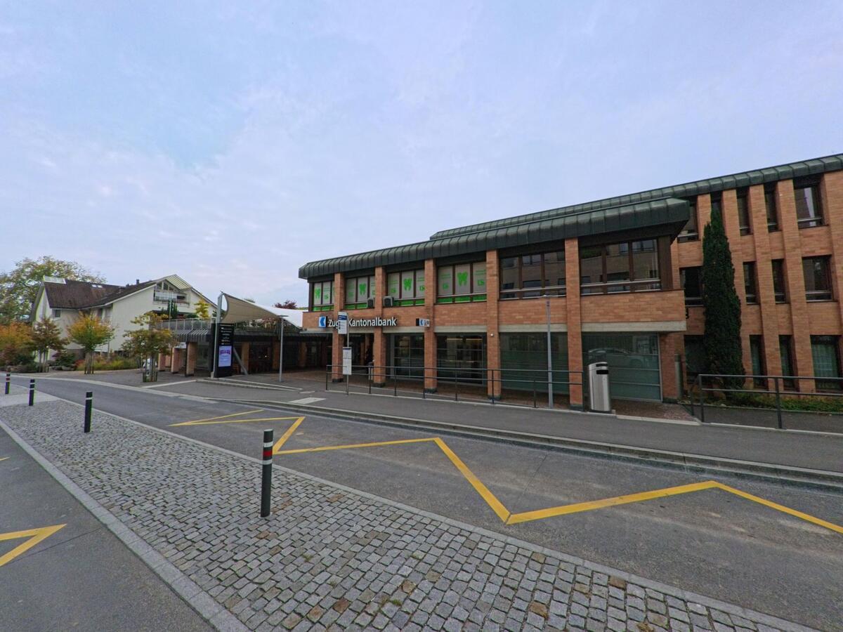 Vor dem Bankgebaeude in Hueenenberg (6331, Kanton Zug) liegt ein gepflasterter Vorplatz mit Markierungen, waehrend das Backsteingebaeude zur Strasse hin ausgerichtet ist.