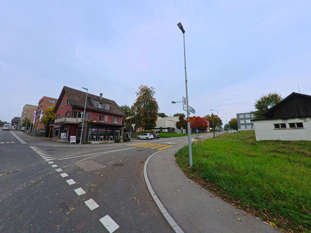 An einer Kreuzung in Hueenenberg (6331, Kanton Zug) steht ein rotes Geschaeftshaus mit Balkon, waehrend die Strasse weiter zwischen Gruenflaechen und Wohngebaeuden verlaeuft.